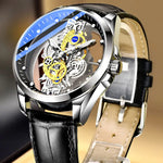 Close-up of a silver skeleton wristwatch with a black leather strap, showing exposed gold and silver gears beneath a clear dial with a blue-and-black outer ring and white hour markers.