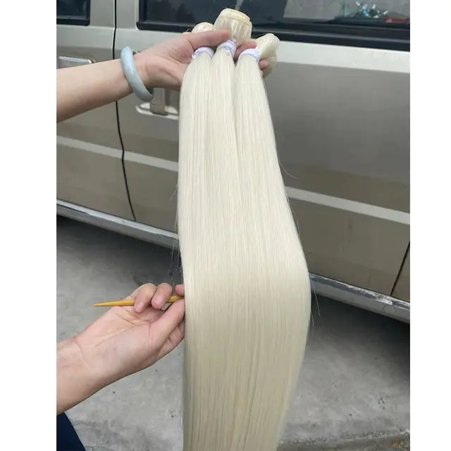 A person holds several long, straight bundles of platinum blonde hair extensions, showing their smooth length and thickness, with a vehicle parked in the background.