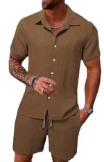 A stylish man with a muscular build is wearing a short-sleeved, button-up green shirt with matching green shorts. He is accessorized with silver rings, a wristwatch, and a layered necklace. His left arm is tattooed, and he is posing casually near a modern poolside setting with a blurred outdoor background.