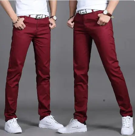 A pair of slim-fit burgundy pants is showcased by a model in two poses against a neutral gray background. The pants are paired with a white belt featuring a bold black buckle design, a white tucked-in shirt, and white sneakers. The model wears a wristwatch and bracelets, adding a stylish touch. The pants have a sleek, modern fit, emphasizing a trendy and casual yet polished look.