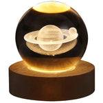Glowing crystal ball lamp on a wooden base with a 3D etched Saturn-like planet and rings inside, illuminated with warm yellow light on a white background.