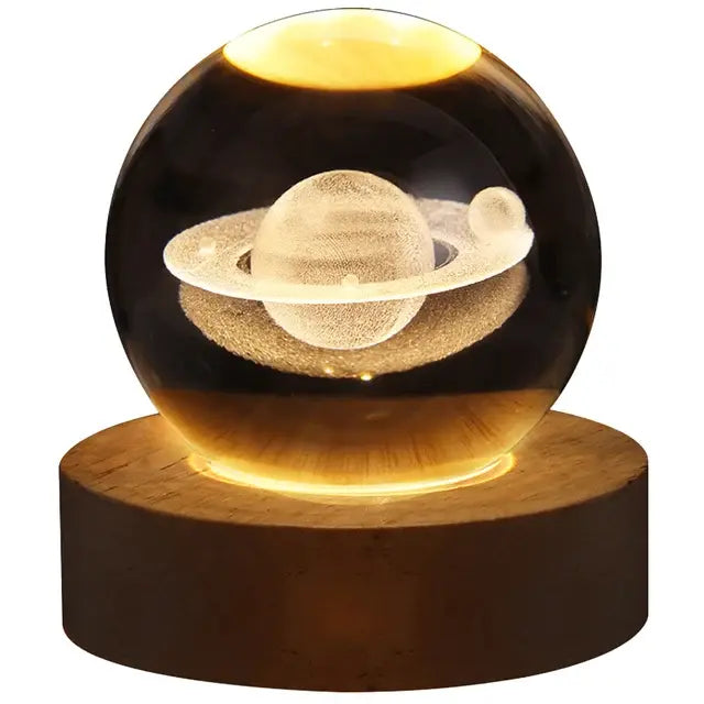 Glowing crystal ball lamp on a wooden base with a 3D etched Saturn-like planet and rings inside, illuminated with warm yellow light on a white background.