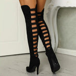 A woman wearing black high-heeled, over-the-knee boots with an open lattice cutout design running up the back, revealing skin between the horizontal straps. She stands indoors on a light wooden floor near a white ornate console table.