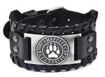 Black braided leather cuff bracelet with snap details and a silver-tone plate featuring a circular emblem with runic-style letters around a paw print, on a white background.