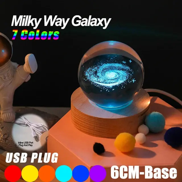 Glass crystal ball night light displaying a glowing Milky Way galaxy on a wooden base, advertised as “7 colors” with a USB plug and 6 cm base, shown with small colorful pom-poms nearby.