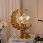 A stylish table lamp with a golden crescent moon shape and a glowing star in the center. The lamp's base is decorated with intricate patterns. The light from the star gives a warm glow, creating a cozy ambiance on the desk, which is topped with a book and a vintage alarm clock.