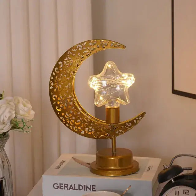 A stylish table lamp with a golden crescent moon shape and a glowing star in the center. The lamp's base is decorated with intricate patterns. The light from the star gives a warm glow, creating a cozy ambiance on the desk, which is topped with a book and a vintage alarm clock.