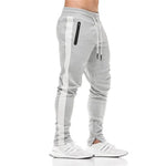 A pair of men's casual fitness workout skinny sweatpants in gray with white side panels, shown from the waist down wearing white sneakers.