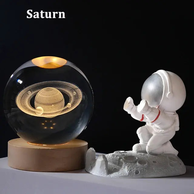A glowing Saturn planet model inside a glass sphere on a wooden base, paired with a small white astronaut figurine on the moon.
