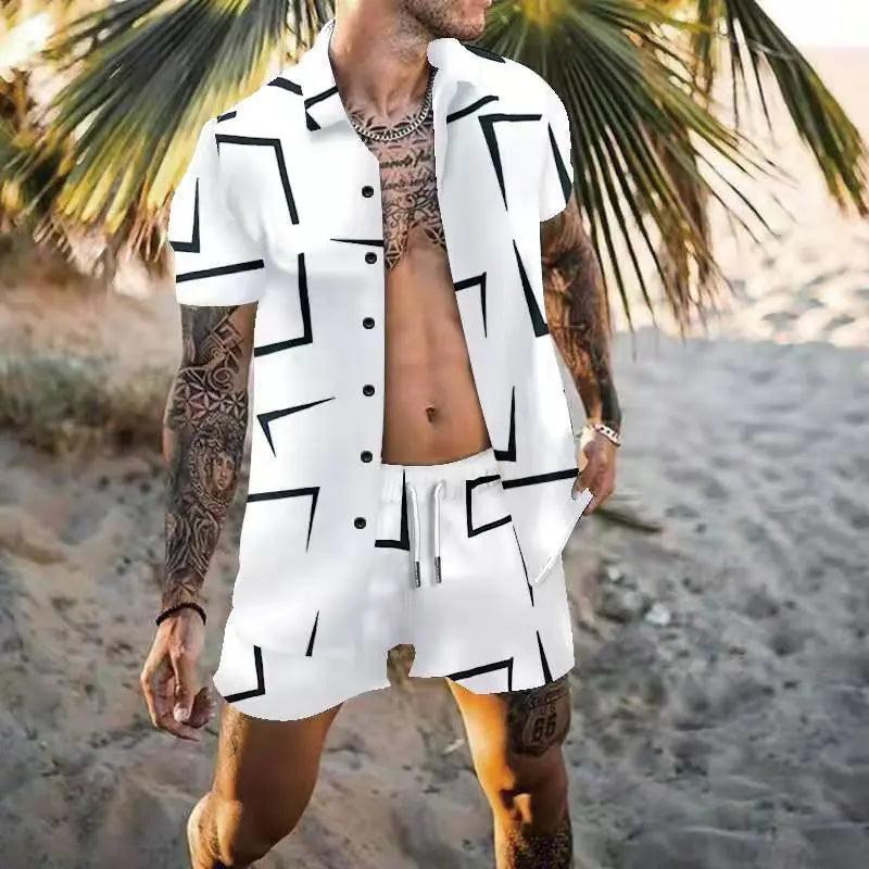 A tattooed man stands on a sandy beach wearing a stylish, matching two-piece summer outfit. The set includes a short-sleeve, button-up shirt and drawstring shorts, both in white with bold black geometric line patterns. He accessorizes with a silver chain necklace and a watch, while palm trees and beach sand provide a tropical backdrop.