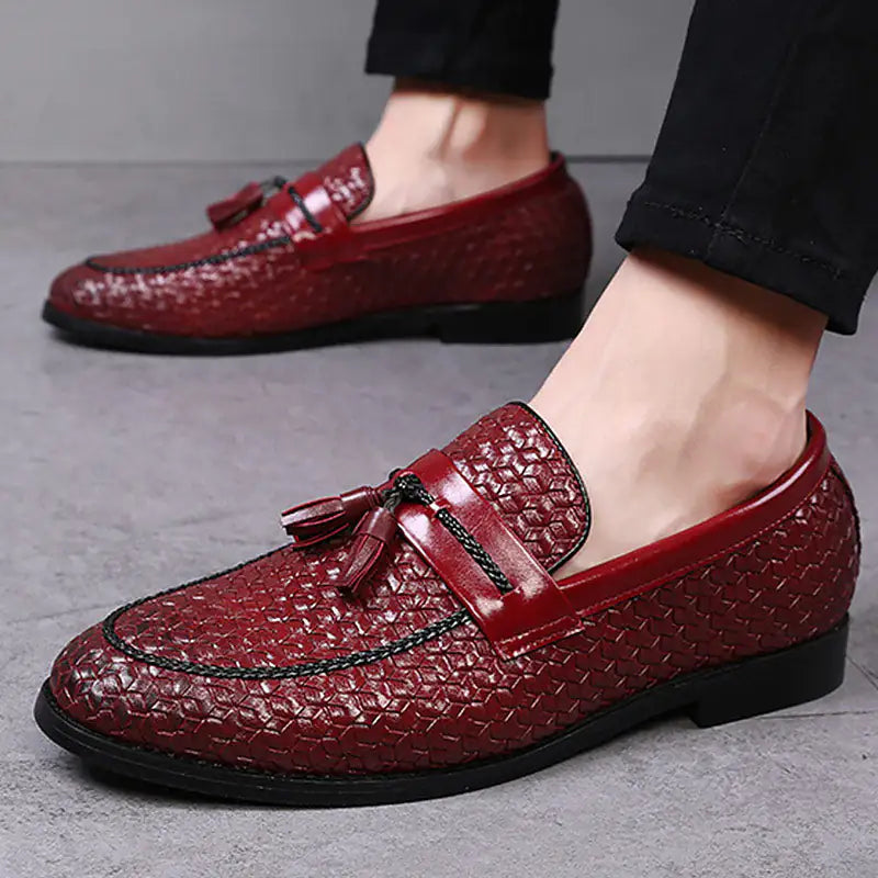 A pair of elegant burgundy leather loafers with a glossy finish. The shoes feature gold-toned metal horsebit detailing across the uppers and are displayed with wooden shoe trees inserted. They have a sleek, elongated silhouette with subtle stitching along the toe cap. The loafers are placed on a dark surface, partially framed by a wooden picture frame, emphasizing their classic and sophisticated style.