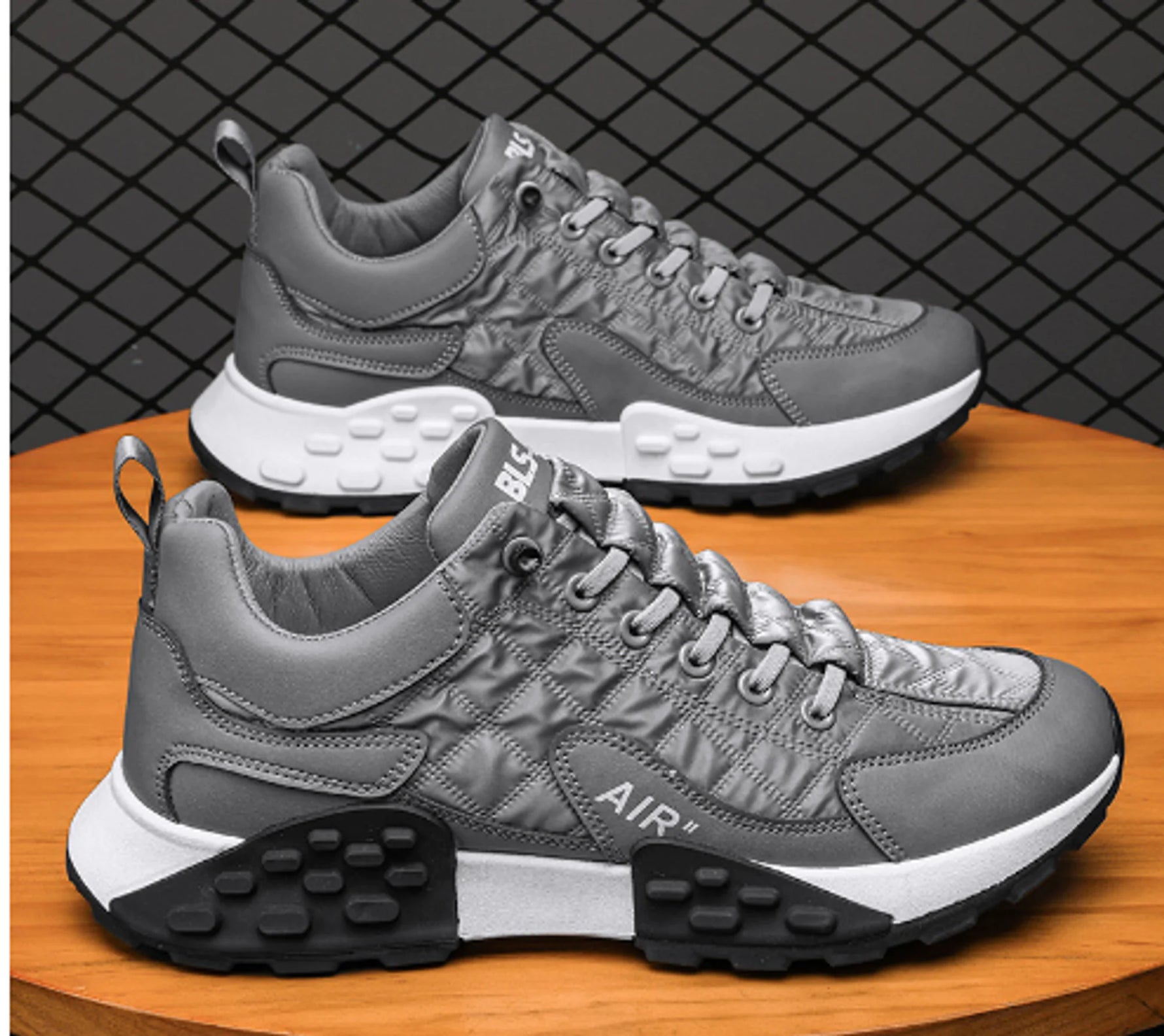 Two gray lace-up sneakers with quilted panels and “AIR” text on the side, featuring white midsoles and chunky black tread, displayed on a wooden surface in front of a chain-link style background.