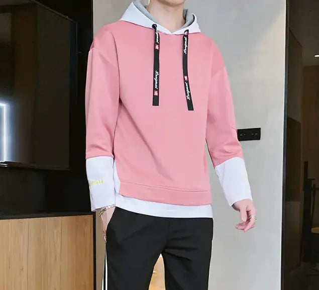 A stylish young man leans casually against a wooden wall while wearing a modern hoodie with black accents. The hoodie features black drawstrings with white text, black cuffs with embroidered details, and a red fabric tag on the left sleeve with 'LONG MA' printed on it. He is also wearing black athletic pants with white stripes on the sides. His accessories include silver rings on his fingers. The background consists of a contemporary indoor setting with a mix of wood and concrete elements.