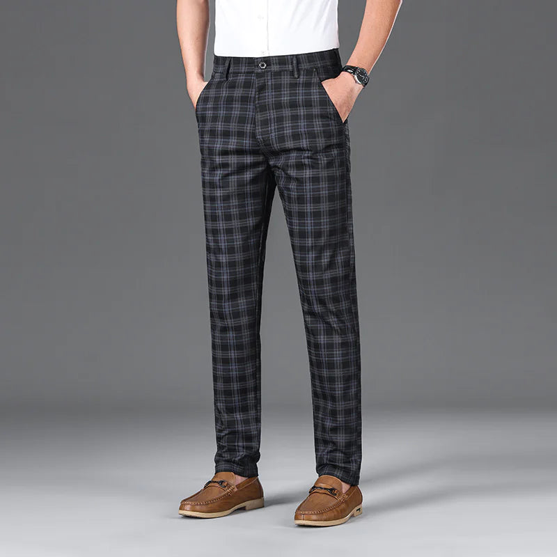A man wearing slim-fit, beige plaid trousers with a black checkered pattern. The trousers have a classic button and zipper closure, belt loops, and side pockets. He pairs them with a white button-up shirt tucked in, brown leather loafers with a decorative metal accent, and a black wristwatch. The background is a simple gray backdrop, emphasizing the stylish and sophisticated outfit.