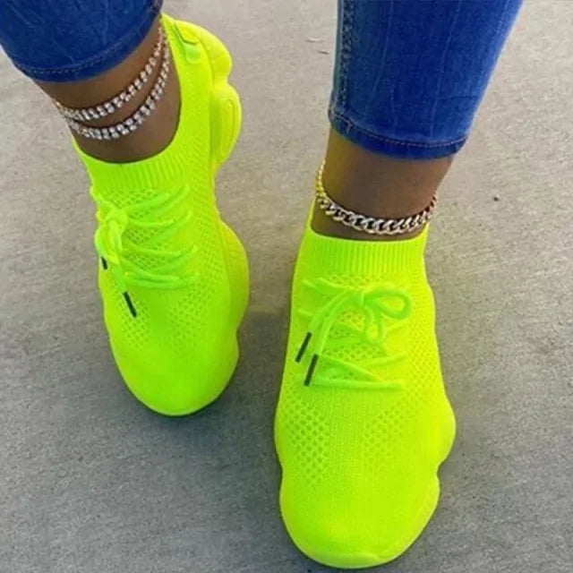 A pair of stylish white knitted athletic sneakers worn by a person standing on an asphalt surface. The shoes feature a breathable mesh design, thick cushioned soles, and gray laces. The person is also wearing white pants and a gold anklet.