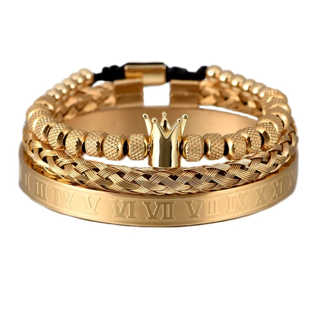 Stack of gold-toned men’s bracelets, including a wide cuff engraved with Roman numerals, a braided chain-style band, and a beaded bracelet with a small crown charm, shown on a white background.