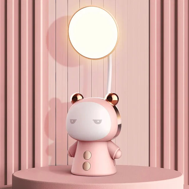 Cute pink bear-shaped desk lamp with a round glowing light on a curved stem, sitting on a circular tabletop against a pink striped wall.