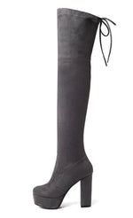 A woman wearing black high-heeled, over-the-knee boots with an open lattice cutout design running up the back, revealing skin between the horizontal straps. She stands indoors on a light wooden floor near a white ornate console table.