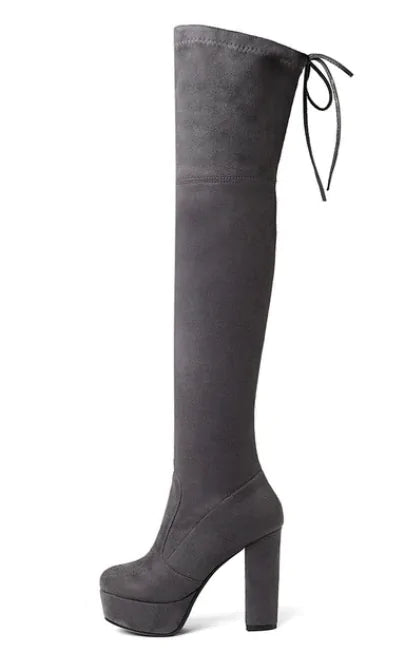 A woman wearing black high-heeled, over-the-knee boots with an open lattice cutout design running up the back, revealing skin between the horizontal straps. She stands indoors on a light wooden floor near a white ornate console table.