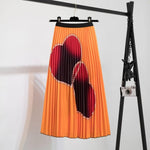 Pleated midi skirt with a gradient design, transitioning from dark black at the waistband to a soft blush pink at the hem, hanging on a black clothing rack next to a vintage-style camera.