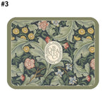 Rectangular decorative mat with a sage green border, vintage botanical floral pattern in muted greens and yellows, and a central oval emblem featuring stylised lettering.