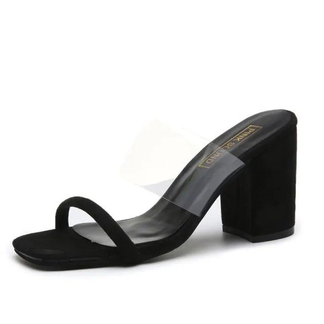 A black slip-on heeled sandal with an open square toe, a single front strap, and a wide clear PVC upper strap. It features a chunky block heel and a sleek minimalist design.
