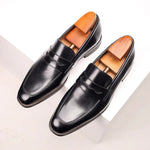 A pair of sleek black leather penny loafers with a polished finish is displayed on a white surface. The shoes feature a classic design with a decorative cutout on the strap and elegant stitching along the edges. Wooden shoe trees are inserted to maintain their shape, adding a touch of sophistication. The overall presentation highlights the refined craftsmanship and luxurious appeal of the loafers
