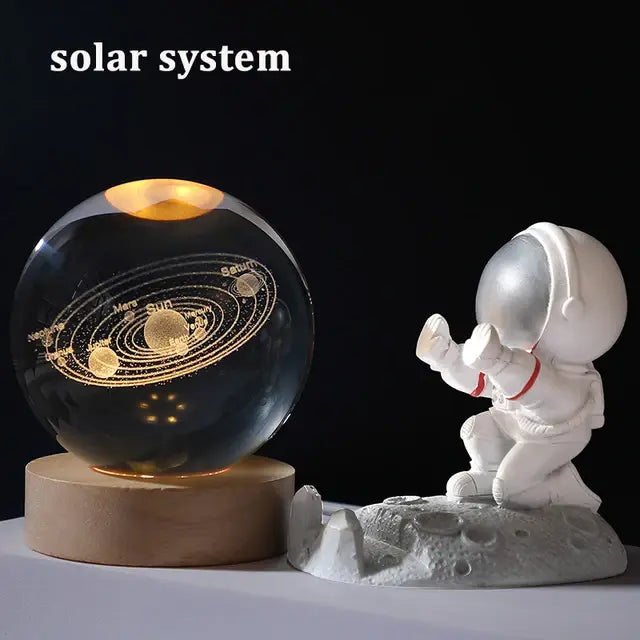 A glowing crystal sphere lamp on a wooden base engraved with a solar system diagram, placed beside a small astronaut figurine kneeling on a moon-like surface, with the words “solar system” at the top