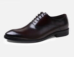 A pair of elegant burgundy leather loafers with a glossy finish. The shoes feature gold-toned metal horsebit detailing across the uppers and are displayed with wooden shoe trees inserted. They have a sleek, elongated silhouette with subtle stitching along the toe cap. The loafers are placed on a dark surface, partially framed by a wooden picture frame, emphasizing their classic and sophisticated style.