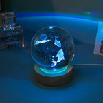 A glowing glass crystal ball lamp on a small round base, lit in blue/teal. Inside the sphere are etched dolphins and tiny star-like specks, creating an underwater/night-sky effect. The scene is photographed in a dim room with other objects softly blurred in the background.