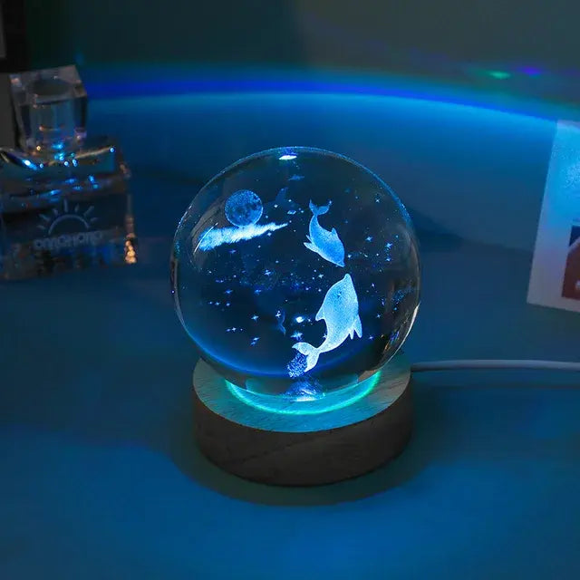 A glowing glass crystal ball lamp on a small round base, lit in blue/teal. Inside the sphere are etched dolphins and tiny star-like specks, creating an underwater/night-sky effect. The scene is photographed in a dim room with other objects softly blurred in the background.