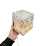 A hand holding a small cube-shaped lamp with a textured glass design, sitting on a wooden base, emitting a warm light. The image has a white background with the lamp slightly blurred at the edges.