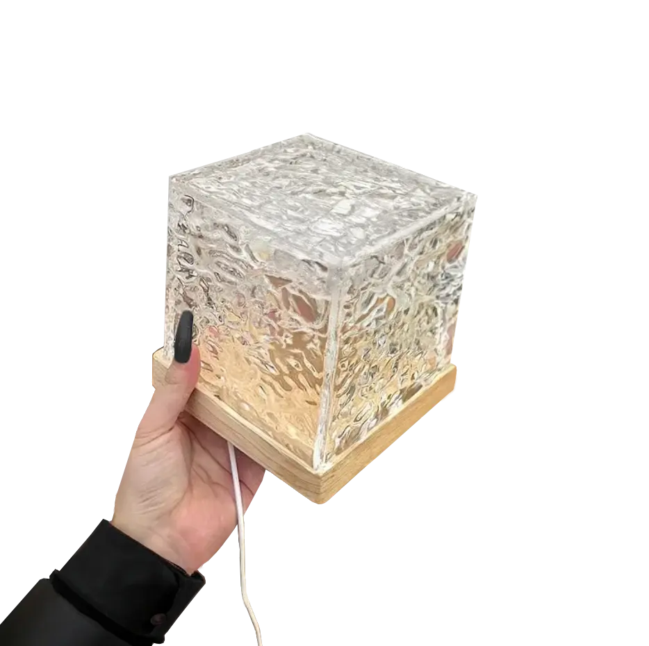 A hand holding a small cube-shaped lamp with a textured glass design, sitting on a wooden base, emitting a warm light. The image has a white background with the lamp slightly blurred at the edges.
