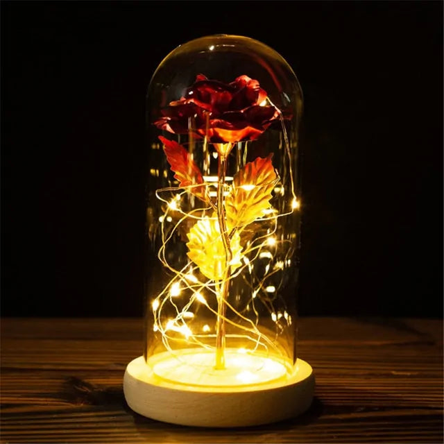 Red artificial rose under a glass dome with warm fairy lights, sitting on a wooden base in a dark setting
