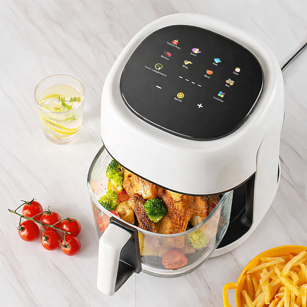 White air fryer with food inside on a light wooden surface, surrounded by vegetables and a glass of water.