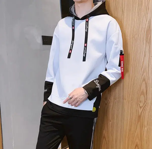 A stylish young man leans casually against a wooden wall while wearing a modern hoodie with black accents. The hoodie features black drawstrings with white text, black cuffs with embroidered details, and a red fabric tag on the left sleeve with 'LONG MA' printed on it. He is also wearing black athletic pants with white stripes on the sides. His accessories include silver rings on his fingers. The background consists of a contemporary indoor setting with a mix of wood and concrete elements.