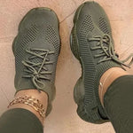 A pair of stylish white knitted athletic sneakers worn by a person standing on an asphalt surface. The shoes feature a breathable mesh design, thick cushioned soles, and gray laces. The person is also wearing white pants and a gold anklet.