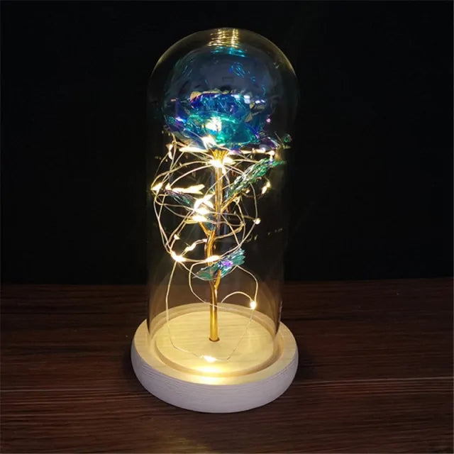 Glass dome display with an iridescent blue rose on a gold stem wrapped in warm white fairy lights, set on a lit base on a dark background.