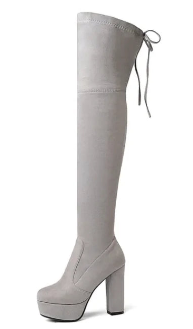 A woman wearing black high-heeled, over-the-knee boots with an open lattice cutout design running up the back, revealing skin between the horizontal straps. She stands indoors on a light wooden floor near a white ornate console table.