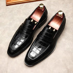 A pair of elegant burgundy leather loafers with a glossy finish. The shoes feature gold-toned metal horsebit detailing across the uppers and are displayed with wooden shoe trees inserted. They have a sleek, elongated silhouette with subtle stitching along the toe cap. The loafers are placed on a dark surface, partially framed by a wooden picture frame, emphasizing their classic and sophisticated style.