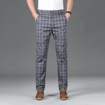 A man wearing slim-fit, beige plaid trousers with a black checkered pattern. The trousers have a classic button and zipper closure, belt loops, and side pockets. He pairs them with a white button-up shirt tucked in, brown leather loafers with a decorative metal accent, and a black wristwatch. The background is a simple gray backdrop, emphasizing the stylish and sophisticated outfit