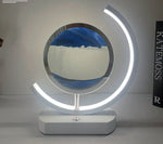 Modern circular tabletop lamp with a white base and two glowing crescent LED arcs surrounding a glass ring filled with blue liquid and white sand, creating a moving sandscape effect.