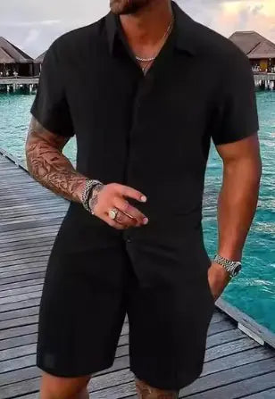 A stylish man with a muscular build is wearing a short-sleeved, button-up green shirt with matching green shorts. He is accessorized with silver rings, a wristwatch, and a layered necklace. His left arm is tattooed, and he is posing casually near a modern poolside setting with a blurred outdoor background.