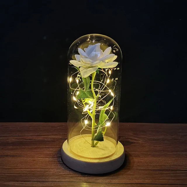 A white artificial rose inside a clear glass dome, wrapped with warm fairy lights, sitting on a round base on a wooden tabletop against a dark background.