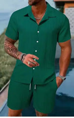 A stylish man with a muscular build is wearing a short-sleeved, button-up green shirt with matching green shorts. He is accessorized with silver rings, a wristwatch, and a layered necklace. His left arm is tattooed, and he is posing casually near a modern poolside setting with a blurred outdoor background.