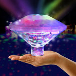 Hand holding a diamond-shaped LED light fixture glowing in shifting rainbow colors, with a dark party-like background filled with colored light spots.