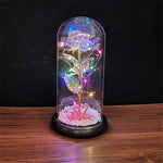 Red rose displayed under a clear glass dome on a wooden base, wrapped with warm white fairy lights and glowing on a rustic wooden surface against a dark background.
