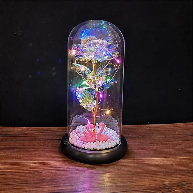 Red rose displayed under a clear glass dome on a wooden base, wrapped with warm white fairy lights and glowing on a rustic wooden surface against a dark background.