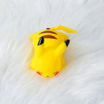 Small yellow Pikachu figurine or toy lying on a white fluffy surface, showing its black-tipped ears, red cheeks, and striped back.