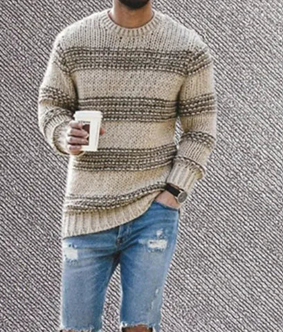 A man wearing a cozy beige and brown striped knitted sweater with a relaxed fit. He is holding a white coffee cup in one hand and has the other hand casually placed in the pocket of his distressed blue denim shorts. The outfit gives off a stylish yet comfortable, casual autumn or winter vibe.